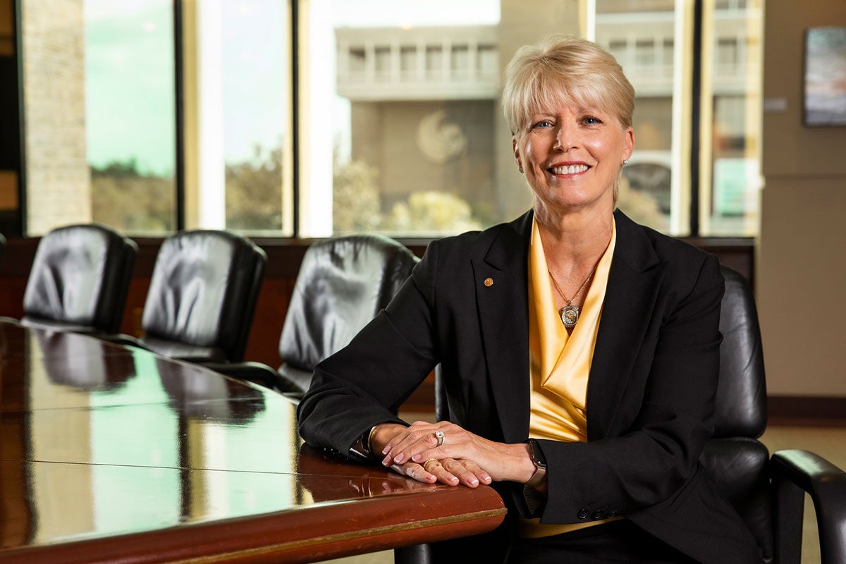 A woman dressed in a black and gold suit sits at a large wood conference table.