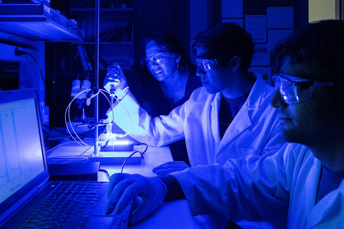 three people in lab coats and protective eyewear sit at a desk with blue light