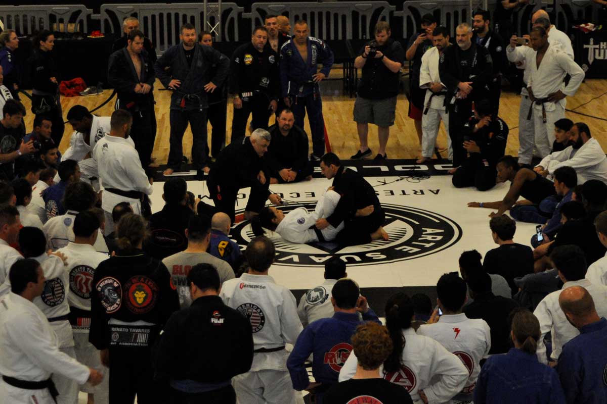 Two men in middle of mat surrounded by circle of onlookers engage in Brazilian Jiu-jitsu