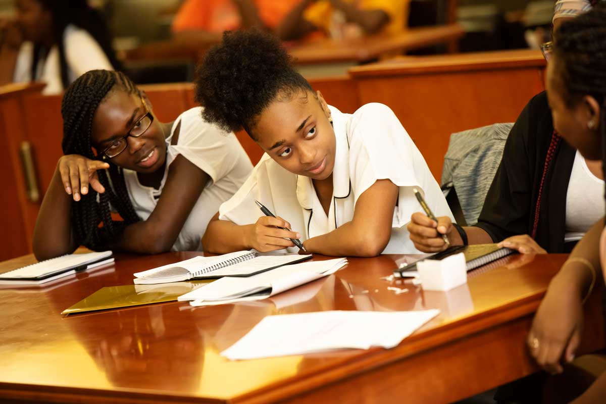 Two teenage girls sit at a wooden desk in a courtroom