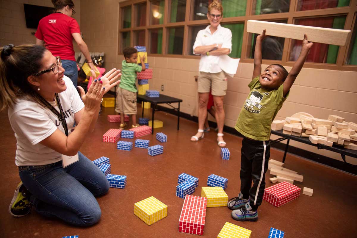 Young boy holds plank of wood overhead while an instructor kneels on the floor and claps for him