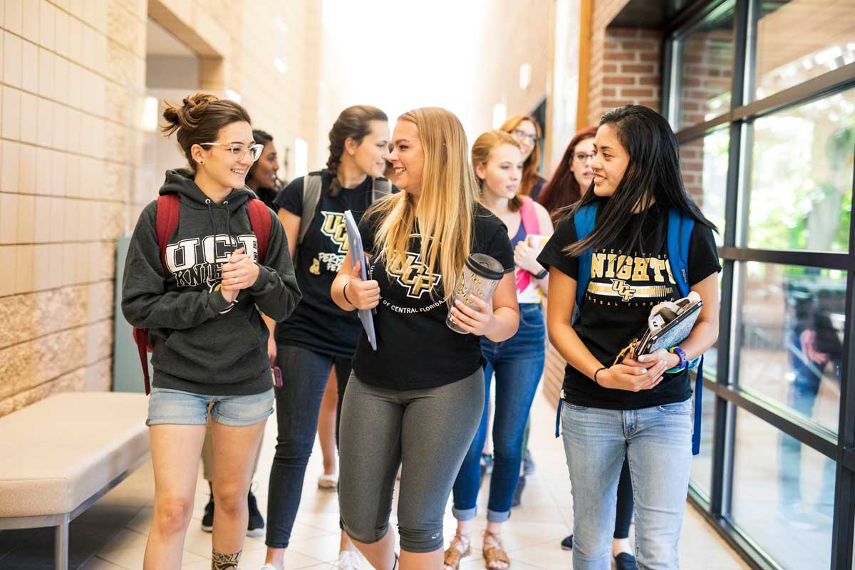 Group of female students wearing UCF shirts and carrying books walk down hallway