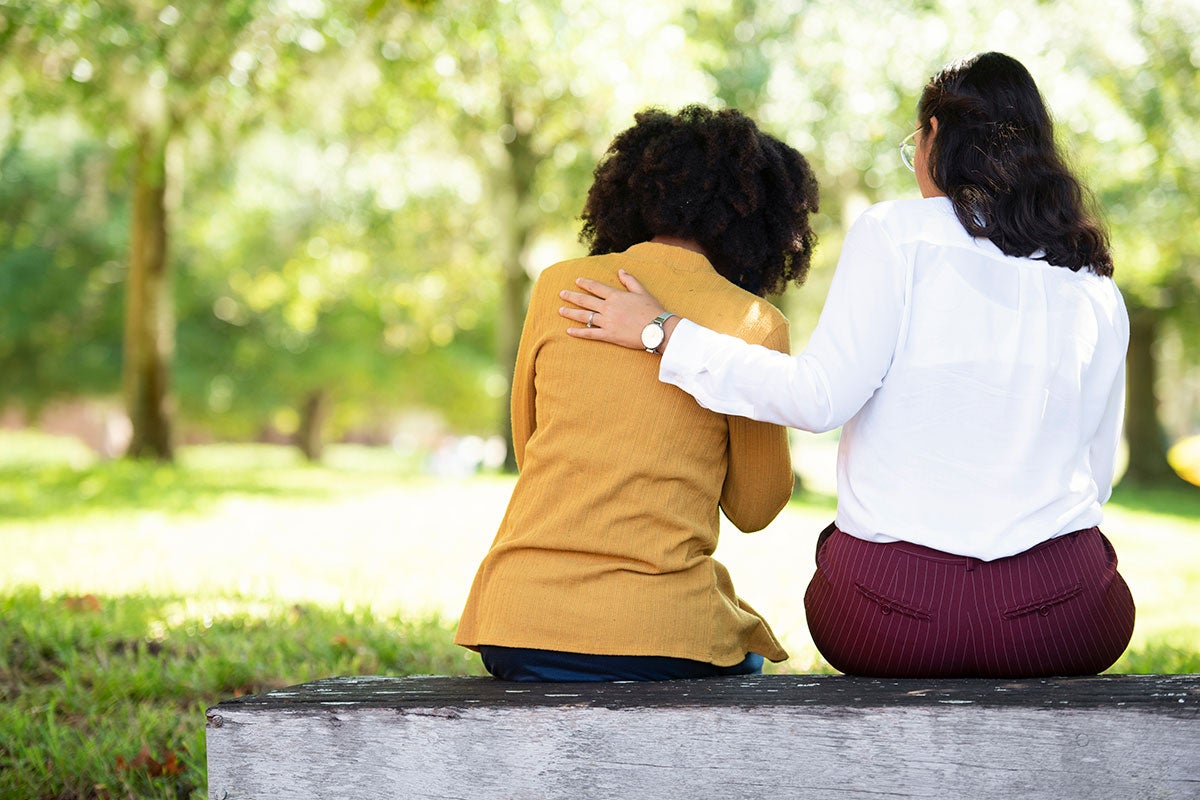 Two women sit on a bench with their back to the viewer. The woman on the right has her hand on the back of the woman next to her, who is holding her face in her hands.