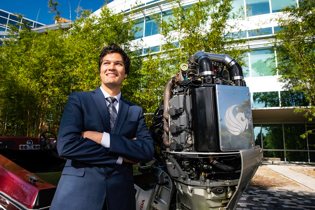 A student stands in a suit with his arms crossed in front of a giant engine on the back of a boat with the UCF Pegasus logo.