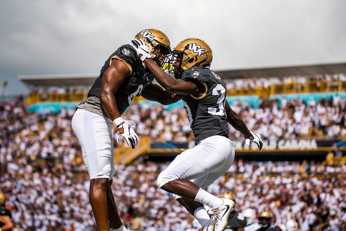 two football players with gold helmets, black jerseys and white pants, embrace in midair with stadium stands in background