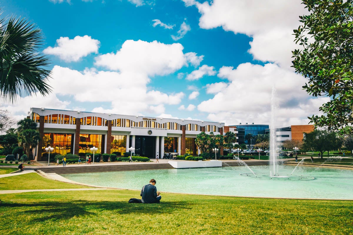 A student sits in front of the Reflecting Pond while facing Millican Hall.