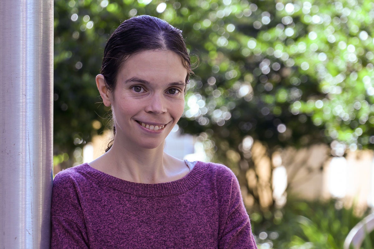 Portrait of a woman wearing a purple sweater while she leans on a wall outside with trees faded in the background