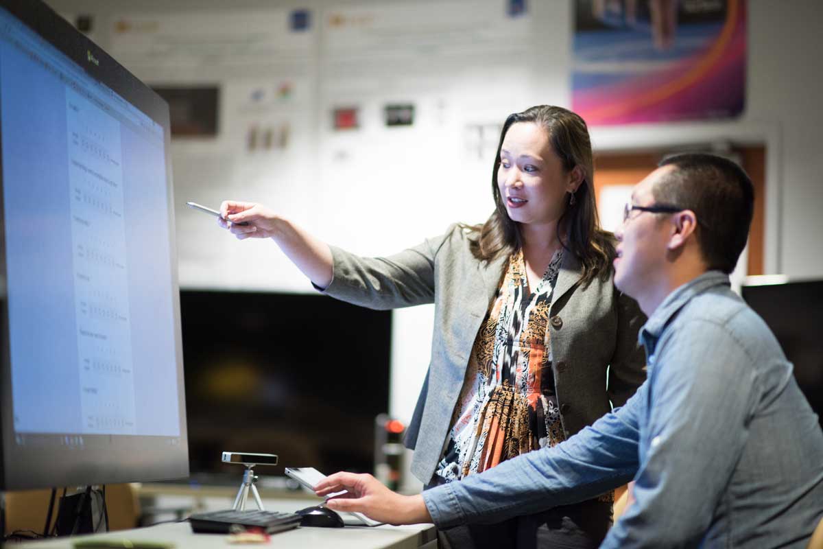 woman standing next to seated man points to large screen