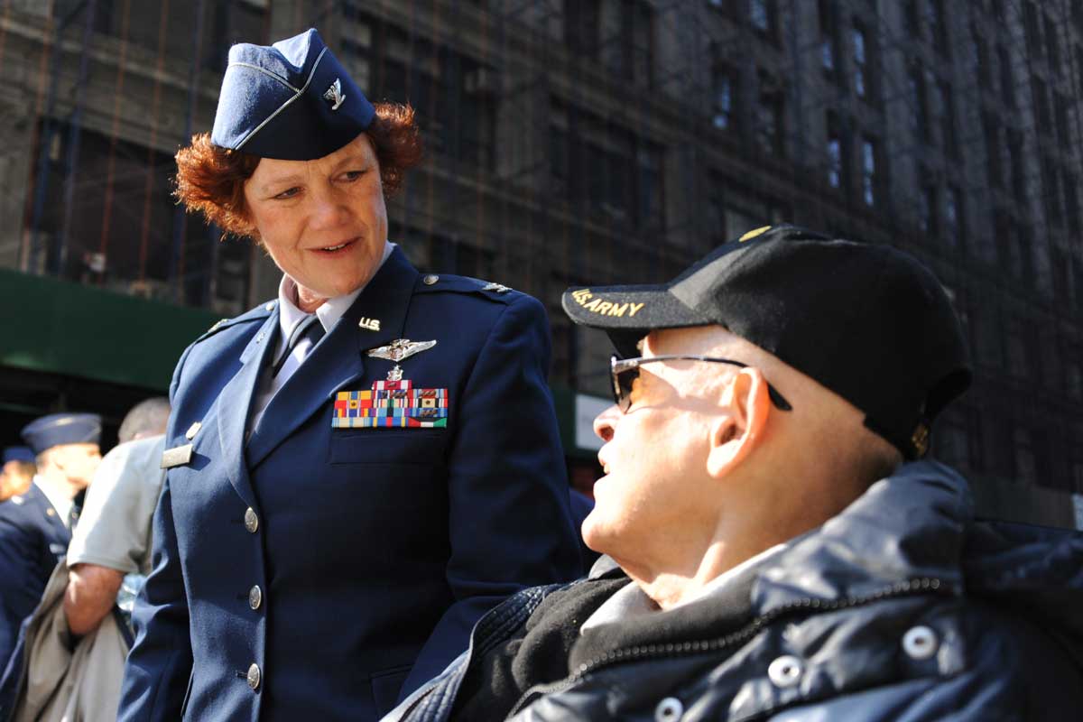 Woman in blue uniform speaks to seated man wearing veteran ballcap