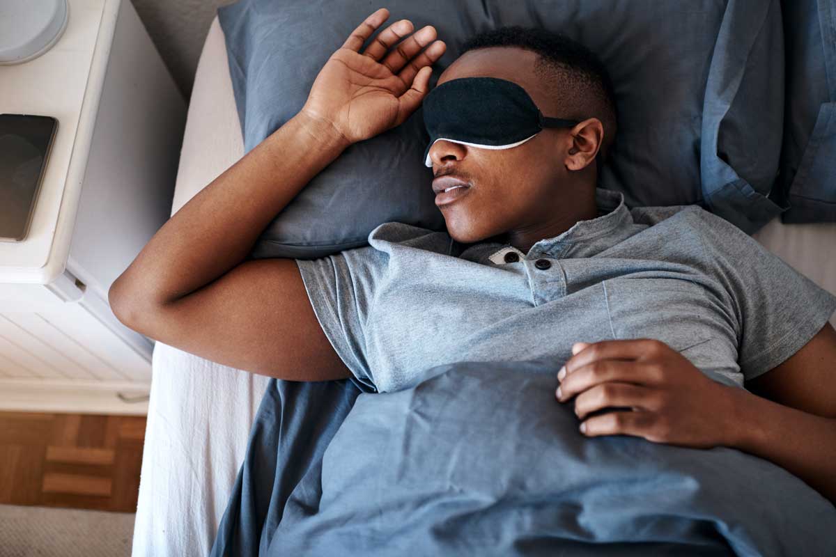 male wearing black eye mask sleeps with his arm relaxed on pillow in bed
