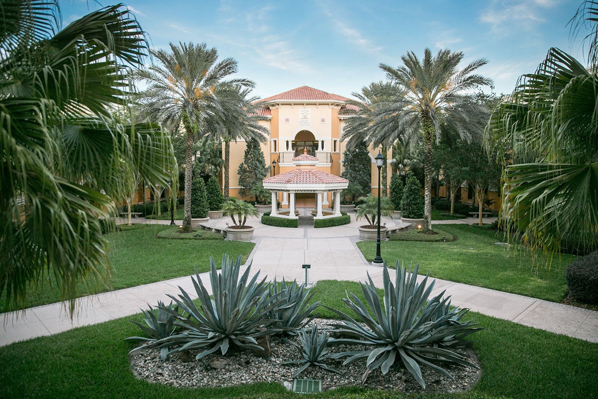 A Spanish-mediterranean-style building is surround by lush green grass and palm trees.