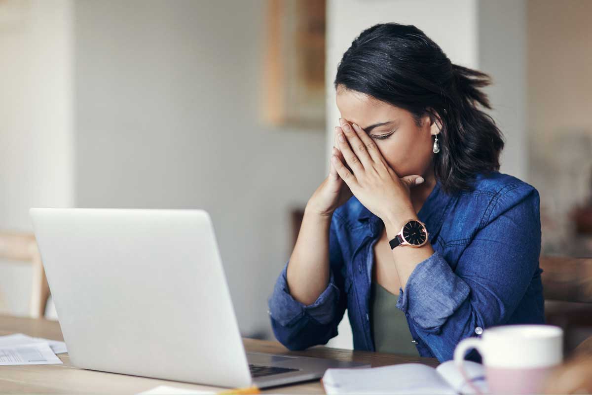 Woman buries face in her hands as she sits at a desk with laptop in front of her