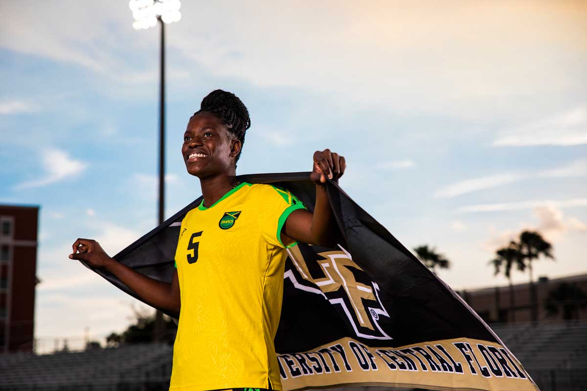 Konya Plummer, wearing yellow Jamaica uniform, drapes UCF flag around her shoulders and stands proudly