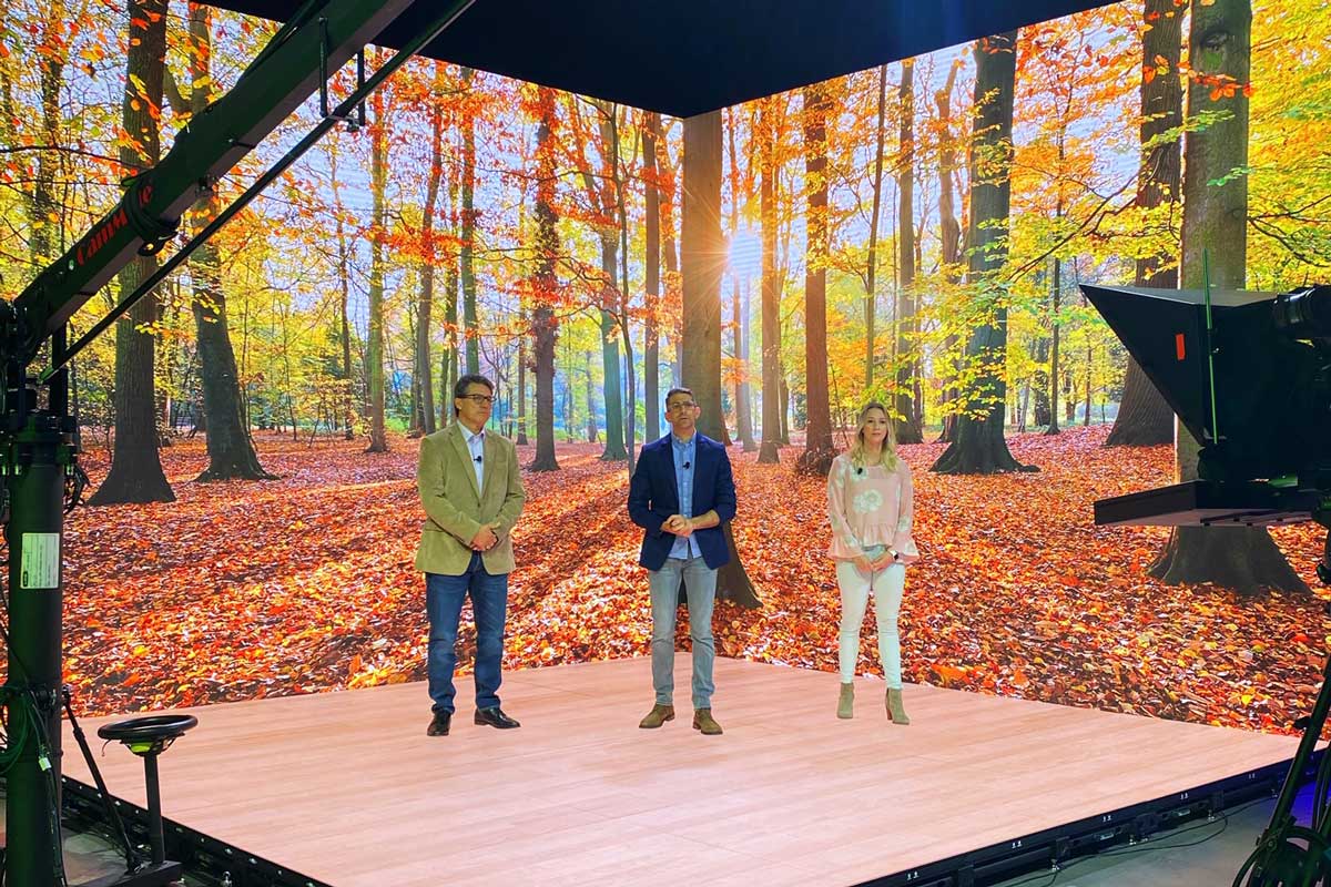 Two men and a woman stand on a studio set with a fall-landscape backdrop behind them