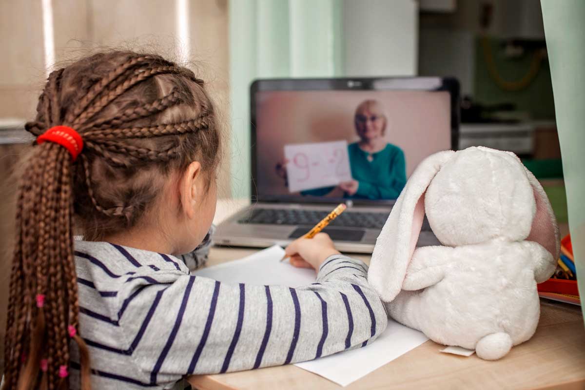 Young girl with braided hair sits at a table while holding a pencil in hand with a laptop in front of her