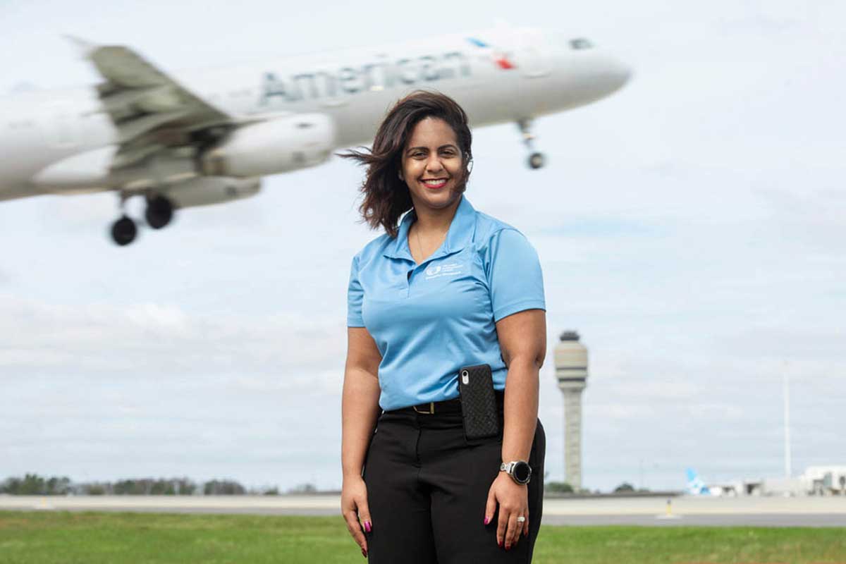 Woman in blue polo shirt stands on airport runway with American Airlines plane taking off in background