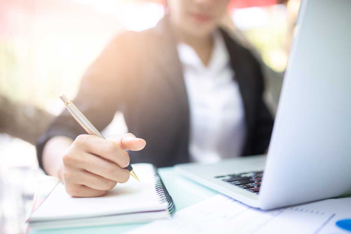 Woman sits in front of open laptop, takes notes