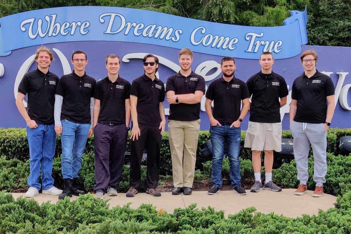 8 members of UCF cyber defense team stand shoulder to shoulder in front of Walt Disney World sign