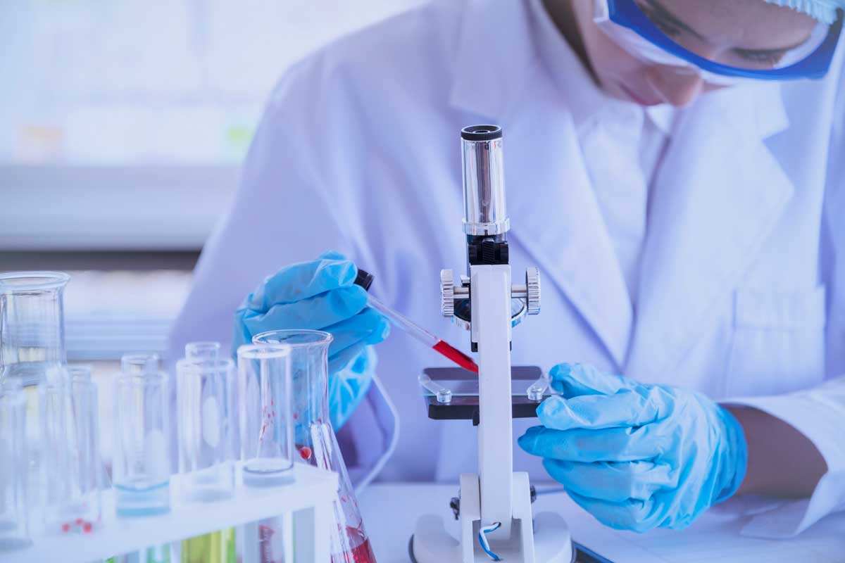 Researcher in lab wearing white coat stands next to microscope