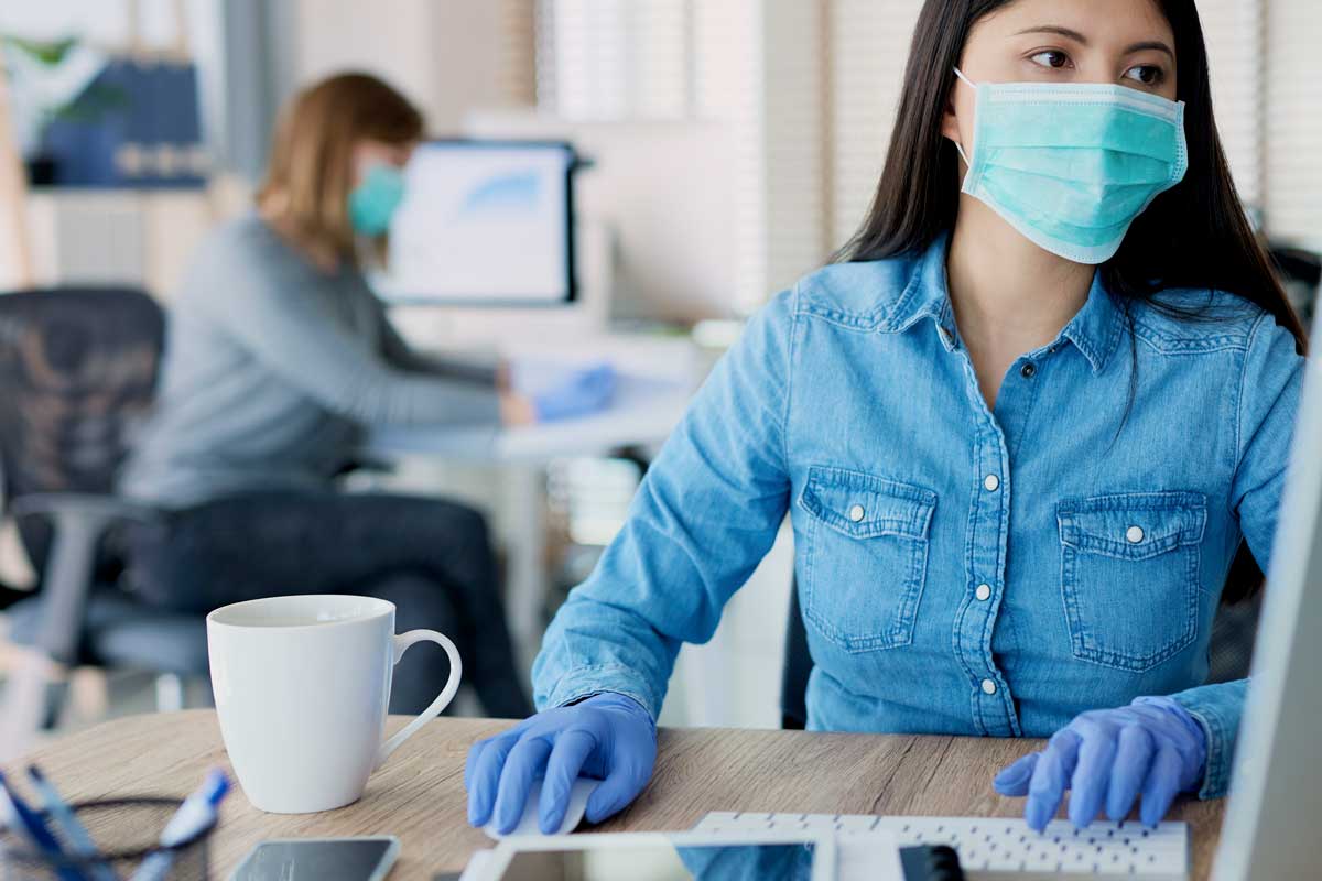 two women wear masks in office at desks with computers