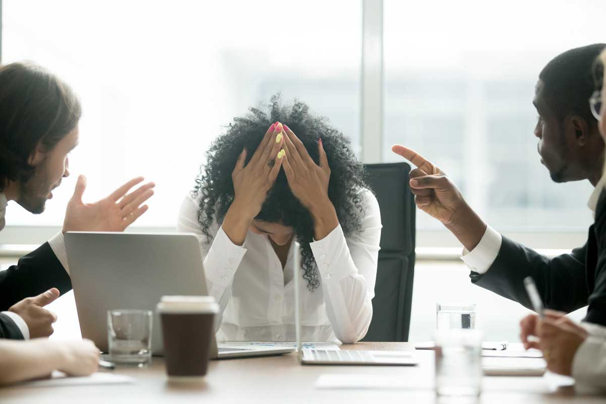 Two men sitting at conference table point at a woman, who has her hands on her head in frustration