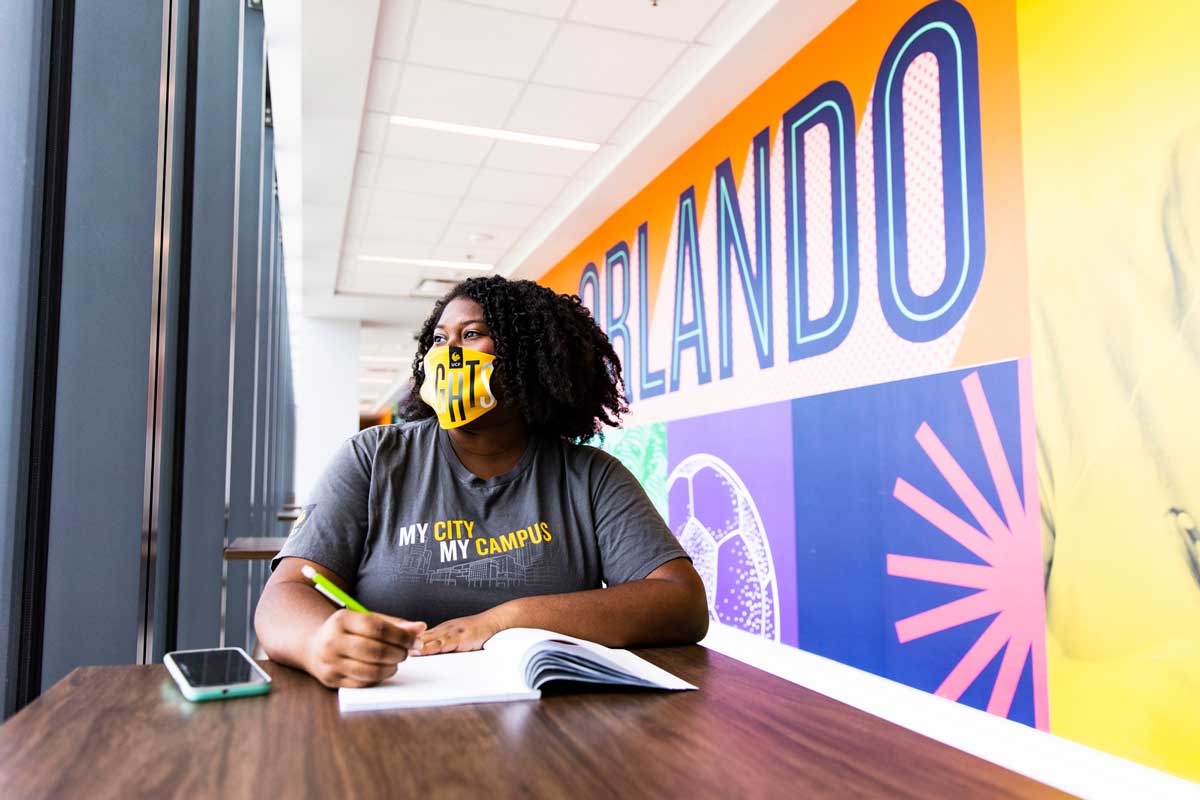 Student wearing yellow face covering sits with an open book in front of a colorful wall