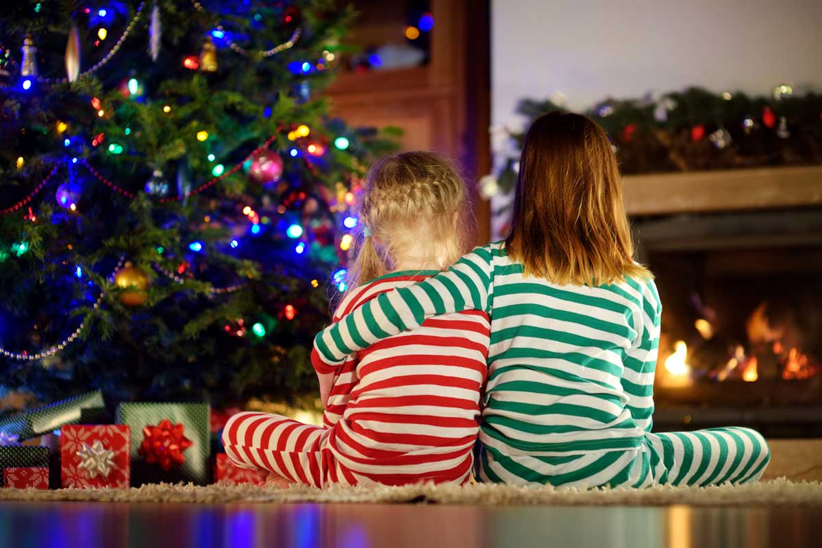 Two young girls in stripped pajamas, one arm around the shoulders of the other, sit by a Christmas tree