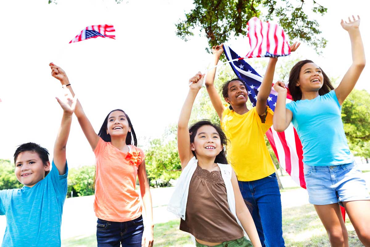 Diverse group of patriotic friends wave American flags at 4th of July parade. The kids are waving the flags and cheering. They are dressed in casual attire.