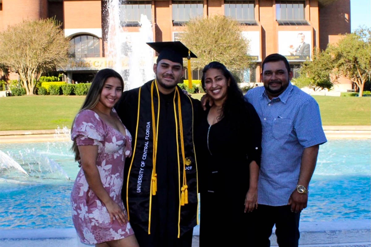 A student wears a cap and gown while posing for a photo with his family.