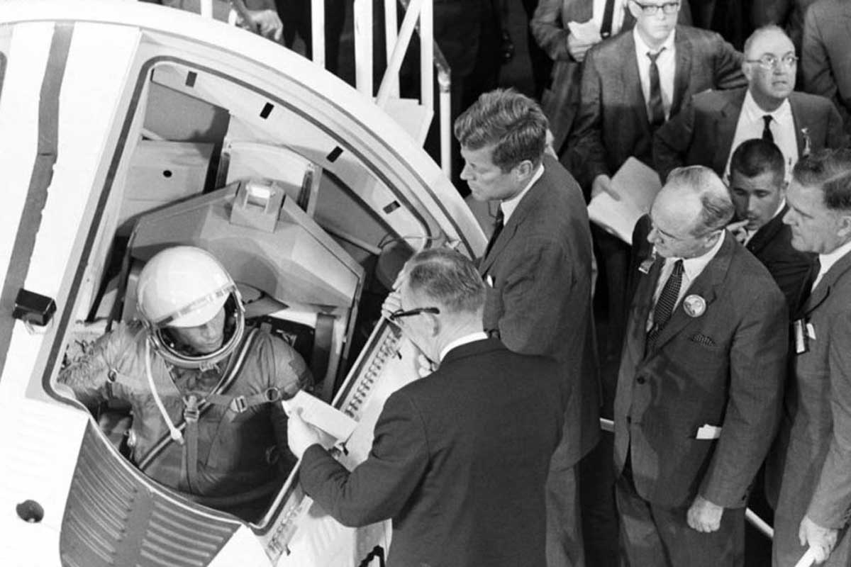 Black and white photo of President John F. Kennedy visiting an astronaut seated in a rocket.