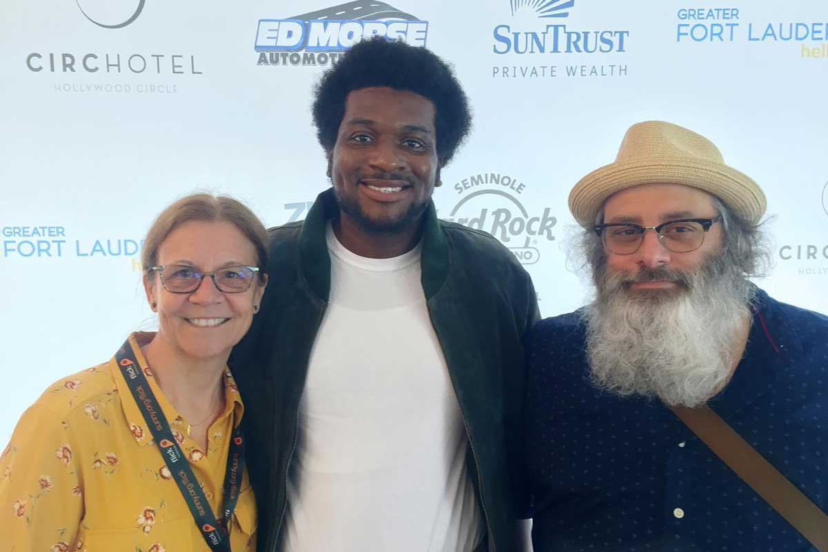 From left to right: woman with glasses and yellow shirt, Black man with green shirt and white undershirt, man with hat and gray beard