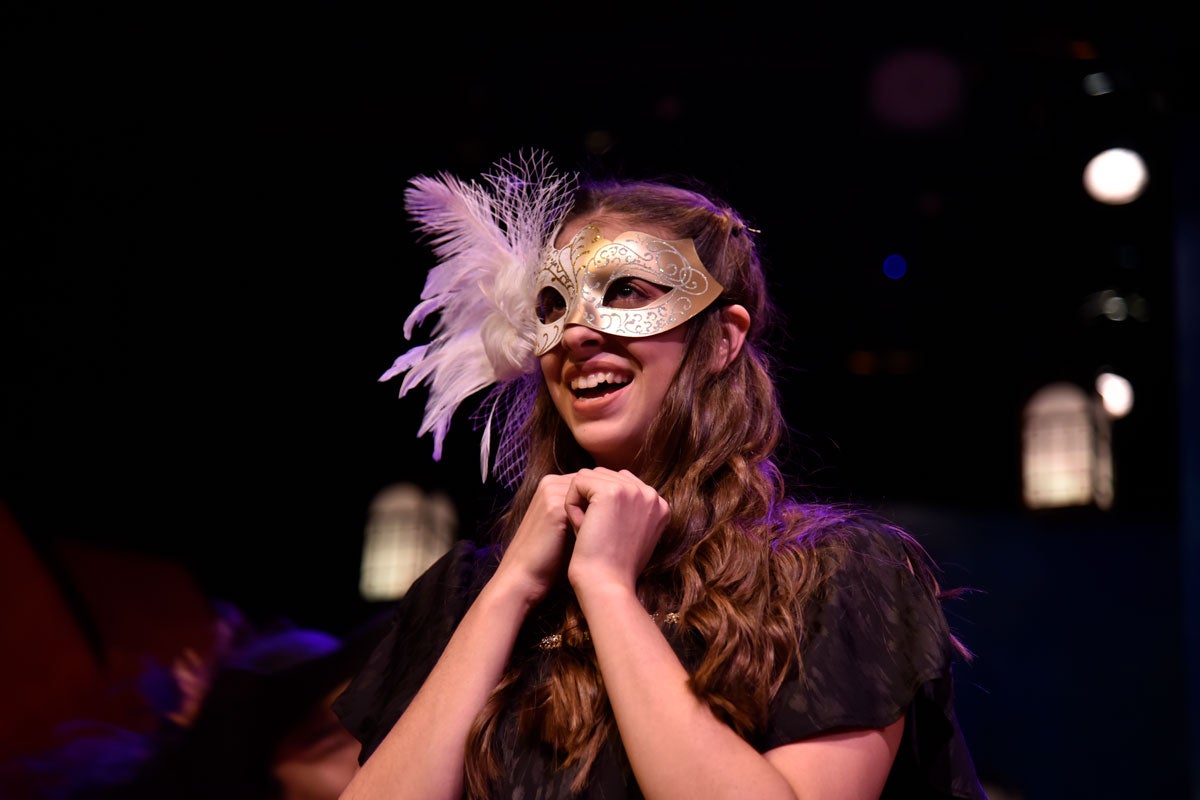 Woman in costume wearing gold mask with feather around the eyes smiles with hands under her chin