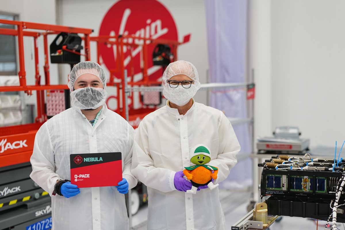 Two men in lab coats and masks stand next to each other, one holds a stuffed animal Citronaut and the other holds a red and black sign that says Nebula