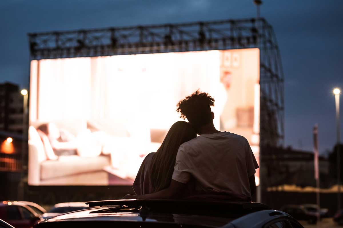 Silhouetted view of attractive young couple, boy and girl embracing, spending time together, sitting in the car while watching a movie in a drive in cinema.