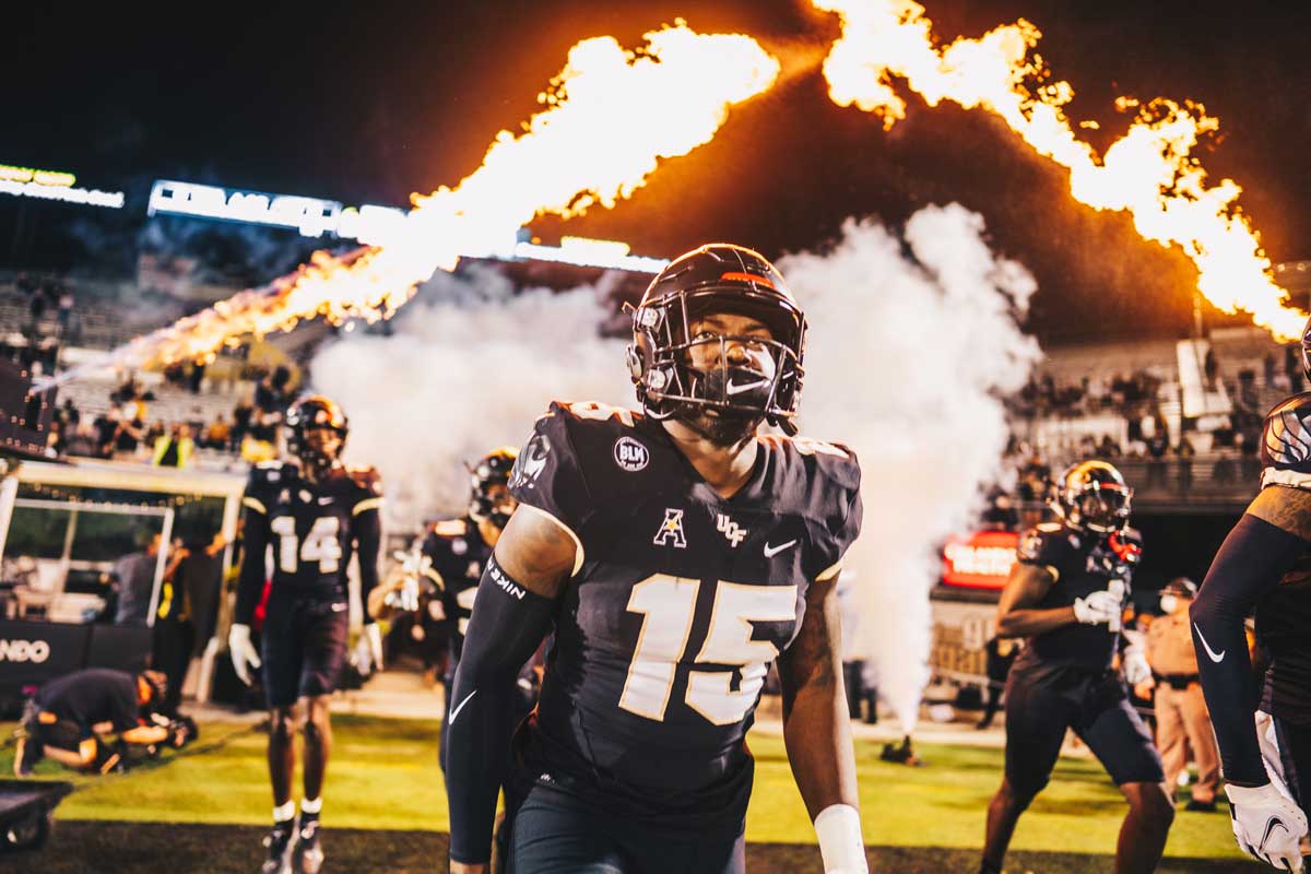UCF football player wearing #15 jersey runs onto the field with pyrotechnics shooting behind him.