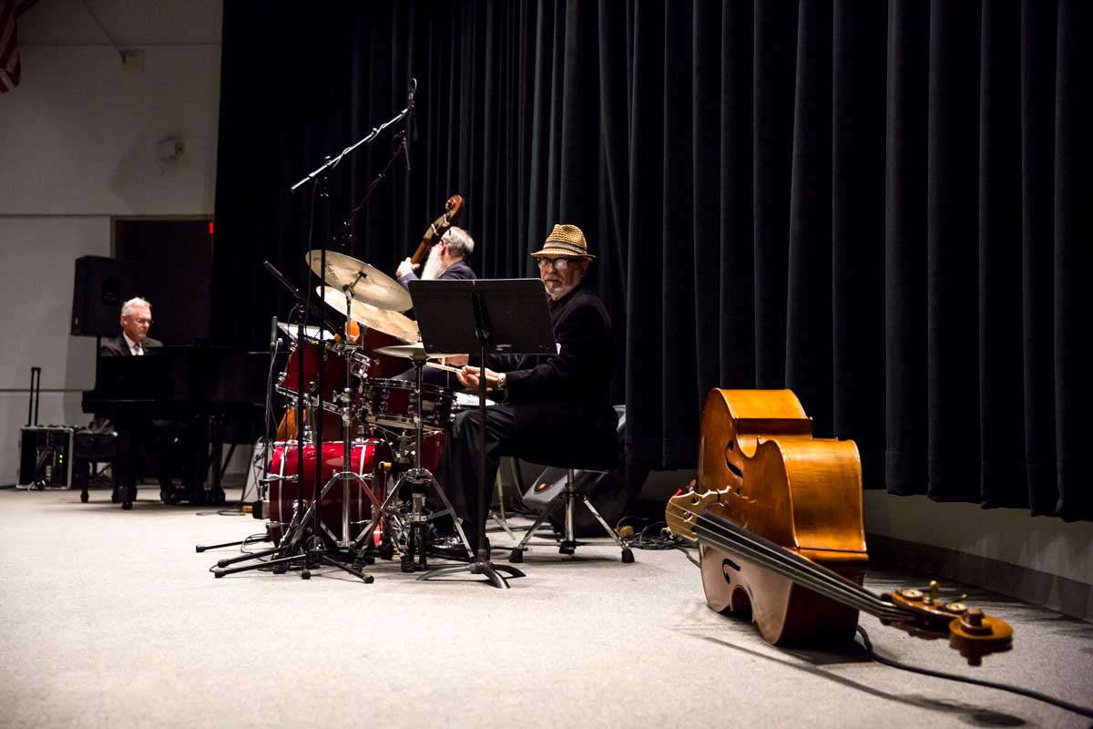 Man sits at a drum set with cello lying on the floor next to him