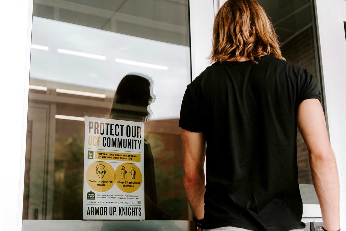Person with shoulder length hair prepares to enter building with a sign on glass door that reads "protect our community"
