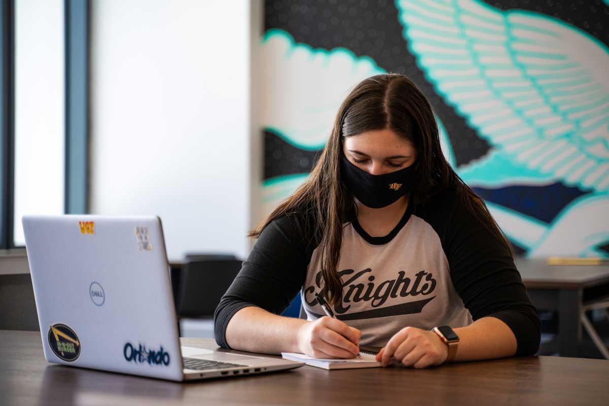 Student wearing black face covering writes on a pad of paper with laptop opened on desk