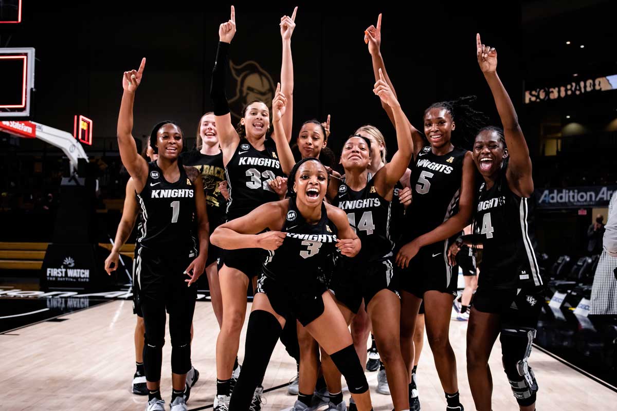 Women's Basketball team huddles in celebration, arms raised and smiling/yelling at camera