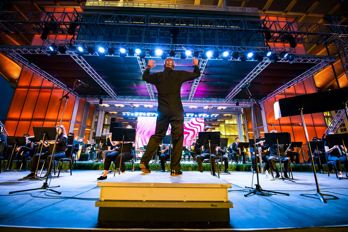 Back view of conductor leading wind ensemble on stage under lights