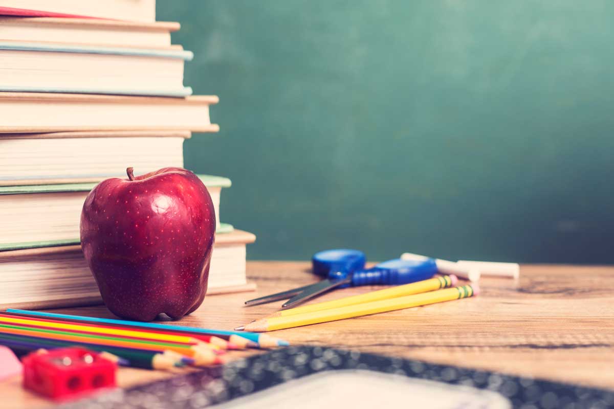 Desk with pencil, workbook, apple, school supplies in front of green chalkboard. Classroom setting.