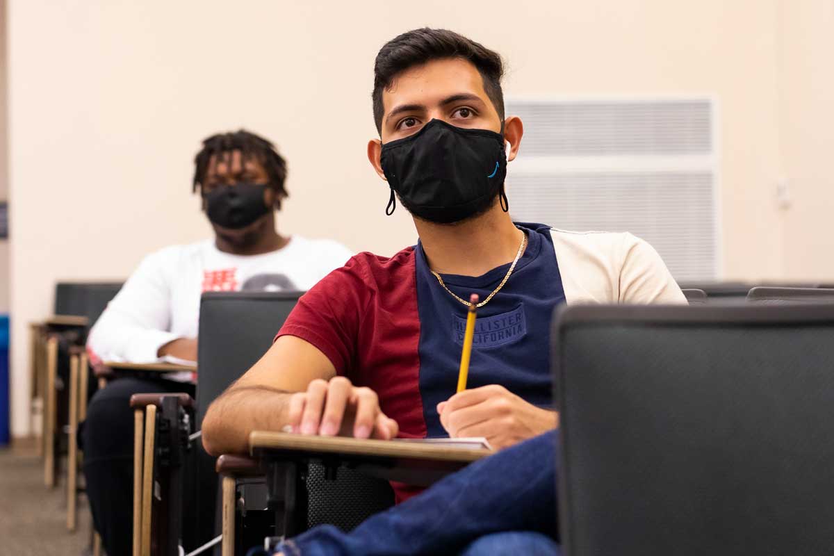 Two students sit at classroom desks while wearing black masks
