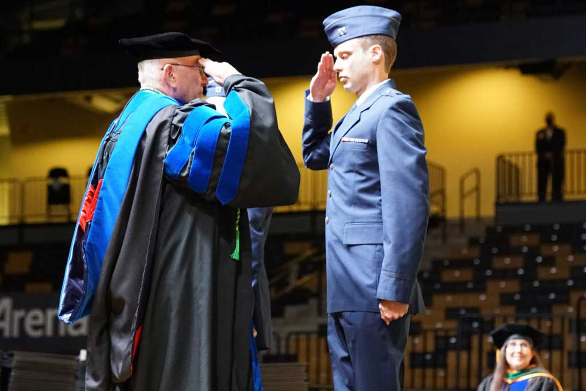 Richard Peppler salutes student in blue military uniform on stage during commencement ceremony