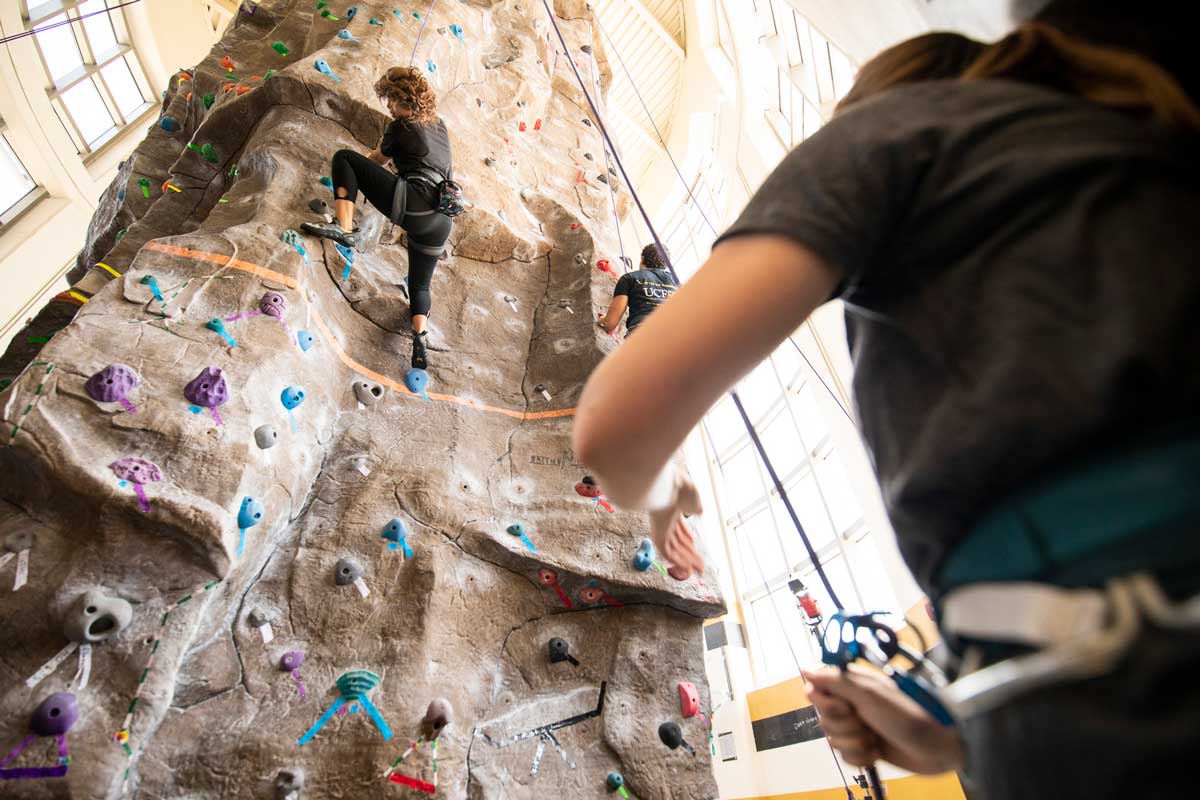 spotter stands on the ground and watches two climbers on rock wall indoors