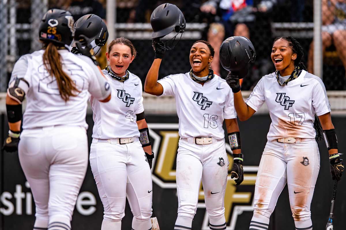 Three softball players raise their helmets in hand to greet their teammate, running home