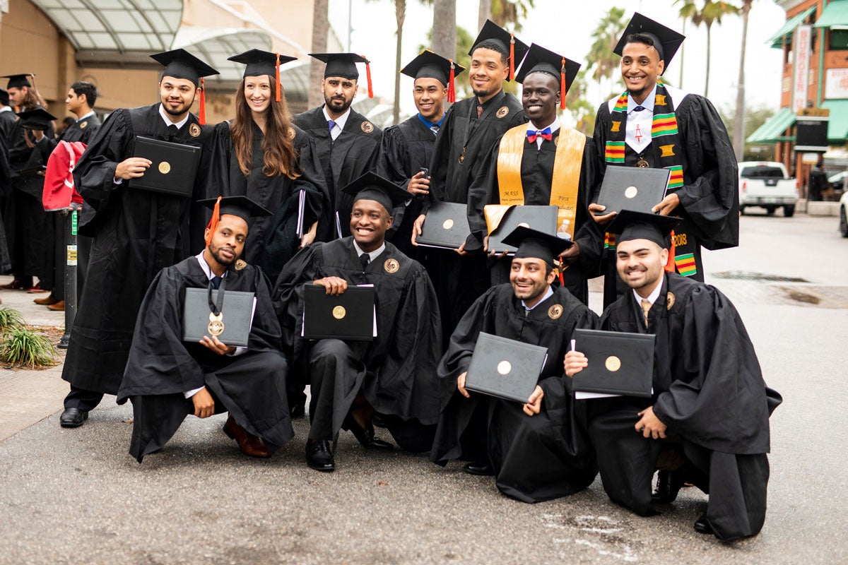 Group of 11 graduates pose with their diplomas in hand outside of Addition Financial Arena