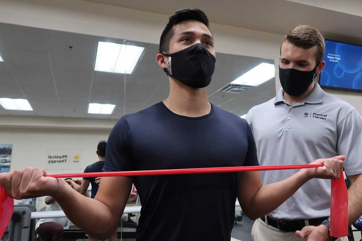 Man stretches red resistance band as a physical therapist observes form