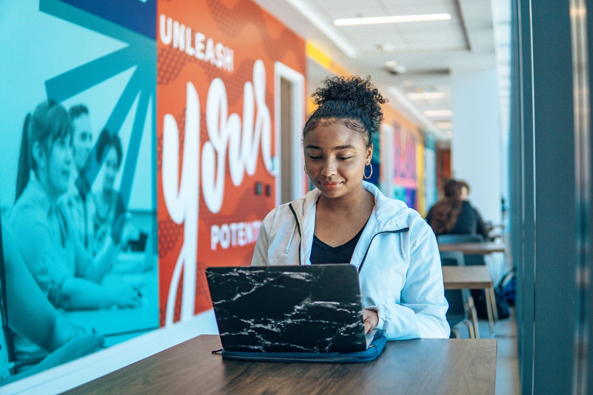 young woman sits with laptop opened in front of her in a hallway