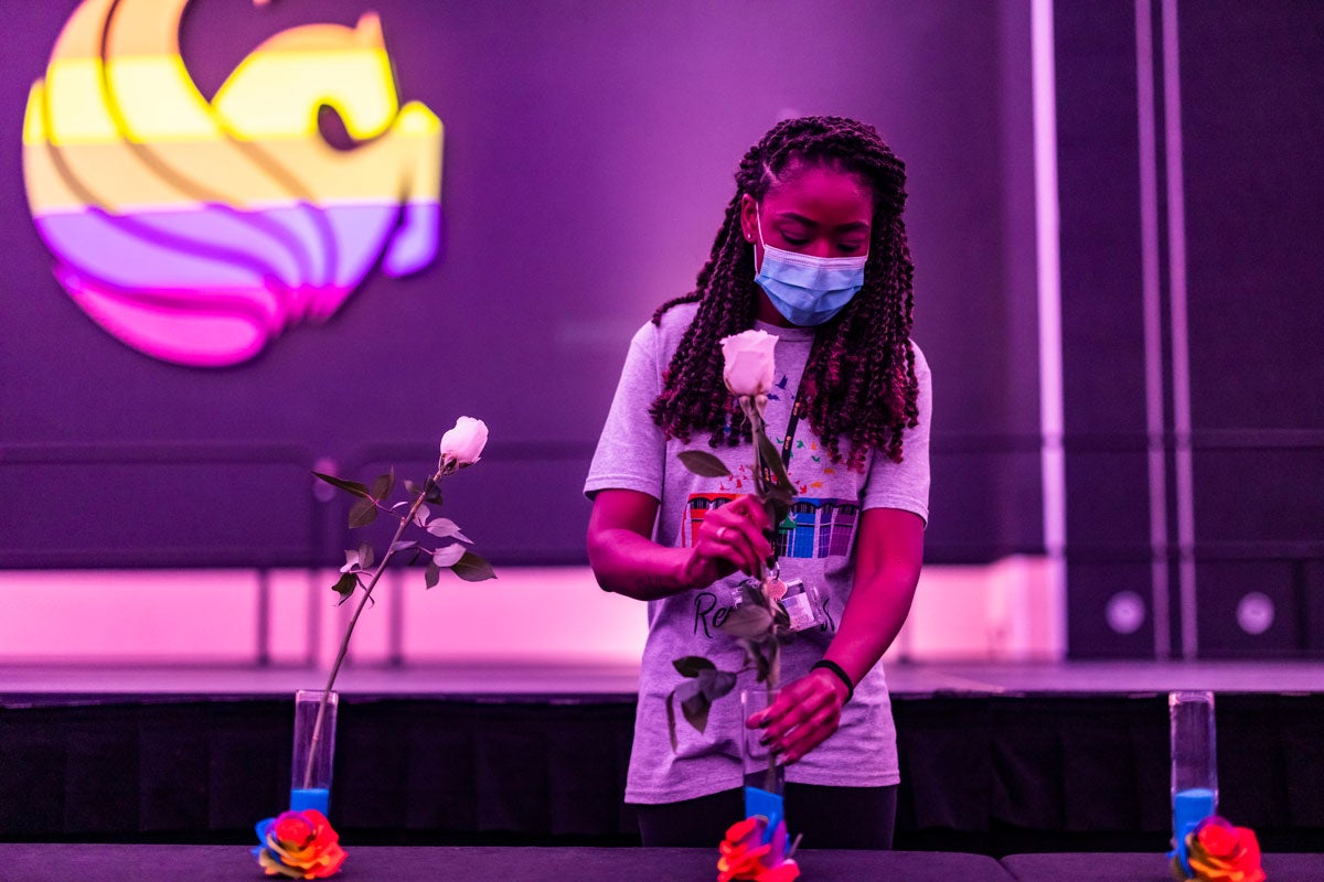Black woman wearing face mask places a rose in a vase with a rainbow Pegasus logo in the background
