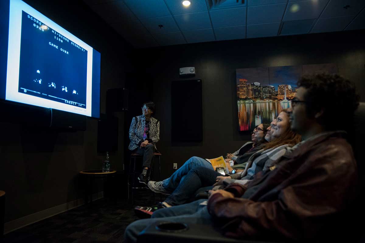 Students sit in a row of seats while observing a screen with a video game message "game over"